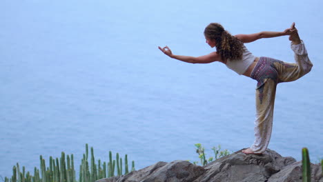 Against-the-backdrop-of-the-deep-blue-ocean-at-sunset,-a-young-woman-performs-yoga-on-a-rocky-seashore,-exemplifying-a-healthy-lifestyle,-harmony,-and-the-intricate-bond-shared-between-humans-and-nature